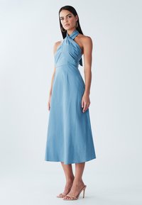 Hele sinine kaelust kinnituv kleit, millel on vormitud ülakeha ja voogav seelik, valmistatud siledast kangast. Omaduseks pööratud esikujundus ja poolpikkus kaltsupikkus.