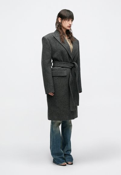 Kvinde iført oversized mørkegrå uldfrakke med bælte over falmede, vidde jeans og åbne sorte sandaler, stående mod en ensfarvet baggrund.