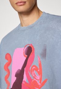 Mand iført en falmet blå T-shirt med abstrakt pink og rød grafisk design på brystet, viser kun hals og nederste del af ansigtet.