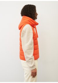 Oranger Puffer-Weste mit hohem Kragen und Kapuze, featuring ein gestepptes Design. Kombiniert mit einem hellbeigen Langarmshirt darunter.