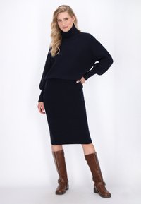 Maglione blu a coste con colletto alto, abbinato a una gonna midi coordinata. Indossato con stivali marroni al ginocchio con un dettaglio a fibbia.