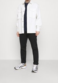 Man med en vit knappad skjorta över en svart t-shirt, svarta uppvikta byxor och silvervita sneakers som står mot en neutral bakgrund.