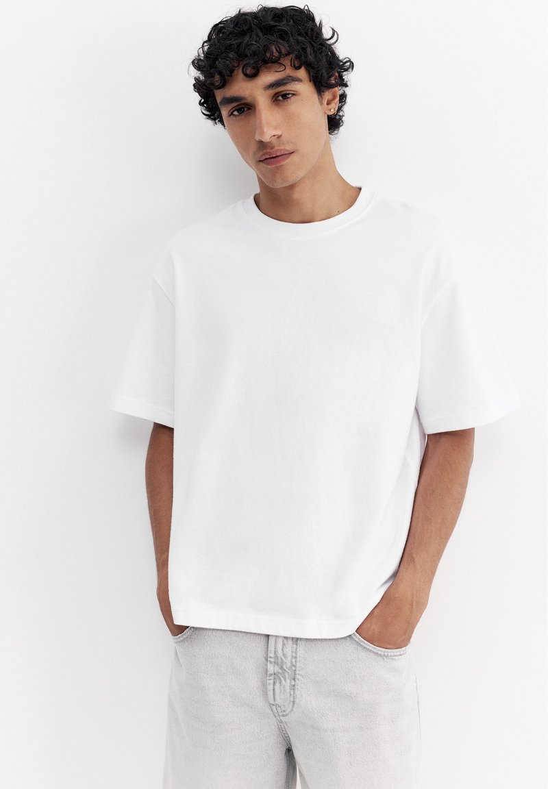 Jeune homme aux cheveux bouclés et foncés portant un t-shirt blanc uni à manches courtes et un jean gris clair, les mains dans les poches, debout contre un mur blanc.