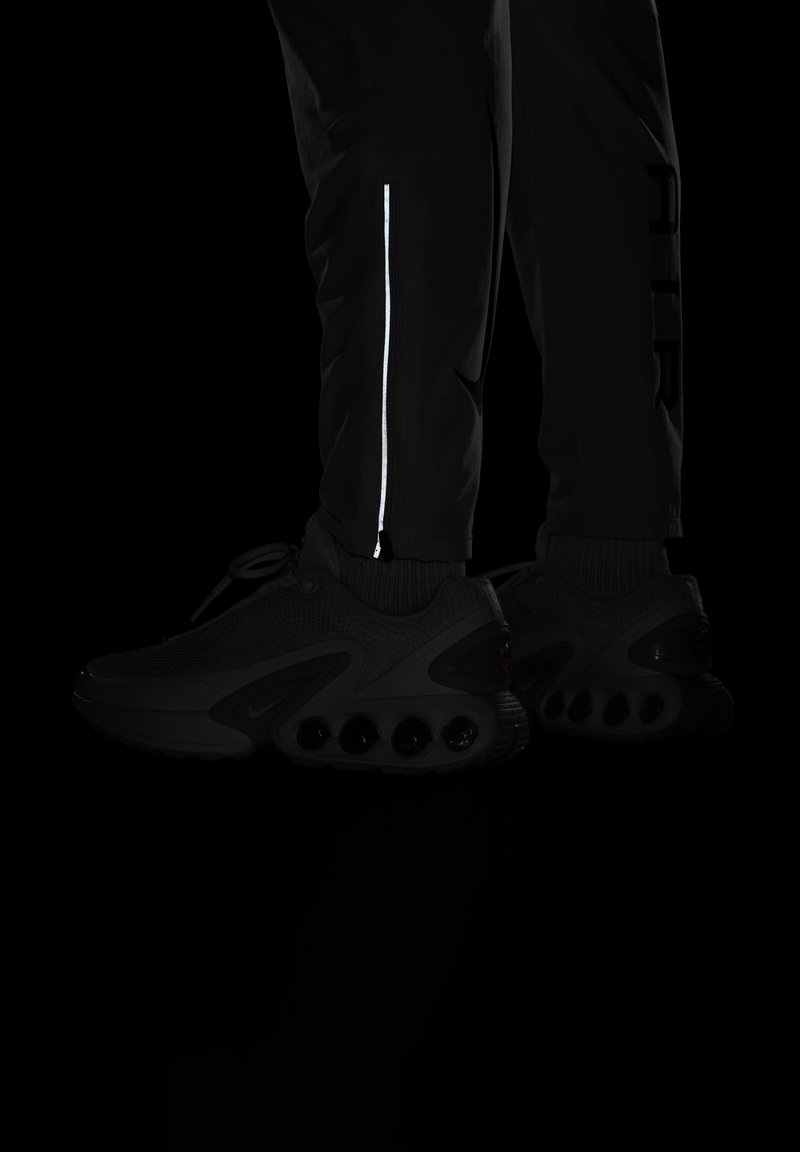 Baskets de sport noires avec un dessus texturé et de grandes unités de coussinage à air. Pantalons noirs ajustés avec un détail de zip réfléchissant.