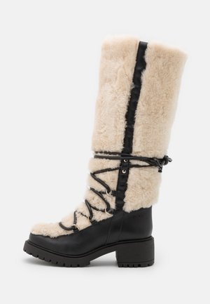 Botte d'hiver haute en cuir noir avec une doublure en shearling de couleur crème, des lacets noirs croisillons sur le devant, et un talon bas épais.