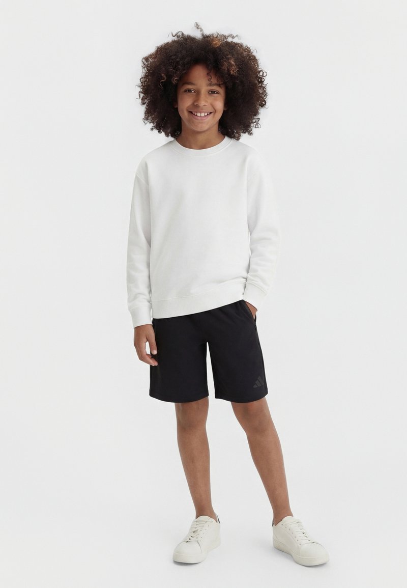 Copil zâmbitor cu părul creț, purtând un tricou alb cu mâneci lungi, pantaloni scurți negri și pantofi sport albi, stând pe fundal alb simplu.