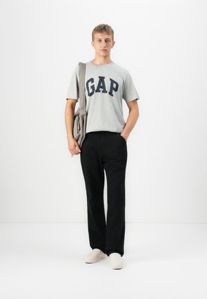 Junger Mann steht da, trägt ein graues GAP-T-Shirt, schwarze Hosen, weiße Schuhe und hat eine graue Umhängetasche auf einem weißen Hintergrund.