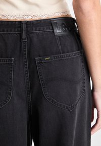 Sorte denimjeans med en sort lærredsetikett med "Lee"-branding i livet, to baklommer med sømdetaljer.