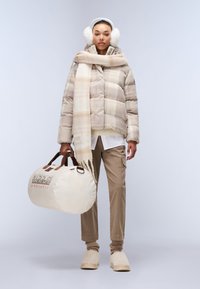 Beige puffjacka med rutmönster, krämfärgad scarf, bruna cargobyxor, beige tofflor och en rund duffelväska med loggdetaljer.