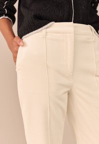Pantaloni in velluto a coste color crema con una texture liscia, caratterizzati da una vestibilità su misura e tasche frontali, abbinati a un maglione scuro.