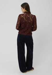 Blouse en dentelle bordeaux avec un col haut et des manches bouffantes, associée à un pantalon large en velours côtelé bleu foncé. Motif floral texturé visible.