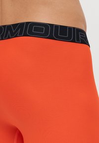 Oransje kompresjonsshorts med en teksturert overflate. Midjebåndet er svart og har merkevarenavnet i fet skrift.