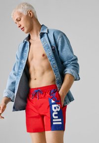 Młody mężczyzna o krótkich blond włosach nosi otwartą kurtkę dżinsową i czerwone kąpielówki z dużym niebiesko-białym logo Red Bull po boku.