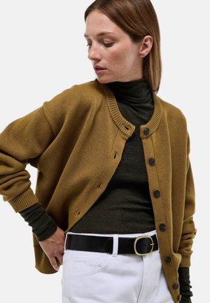 Femme portant un cardigan moutarde marron boutonné sur un col roulé foncé, pantalon blanc et ceinture noire avec boucle dorée, regardant vers le bas.