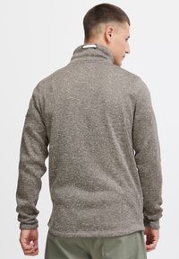 Grå fleece-pullover med et tekstureret mønster, høj krave og lange ærmer, der har en løkke til justering af kravens stramhed bagpå.