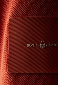 Červená texturovaná tkanina s hladkým obdélníkovým štítkem. Štítek má diagonální proužkovaný vzor a název značky "SAIL RACE" ve stříbrné barvě.