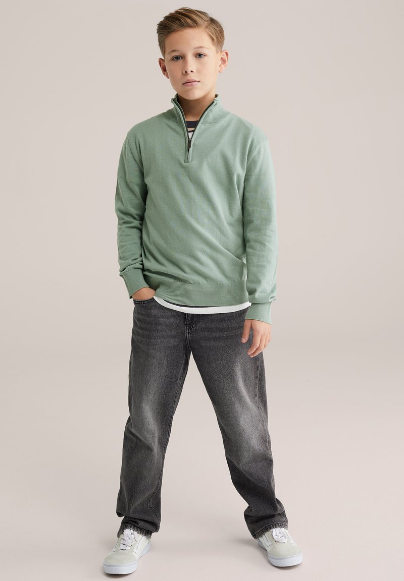 Pull vert clair avec une demi-fermeture éclair, poignets côtelés, porté par-dessus une chemise blanche, associé à un jean gris foncé et des baskets claires.