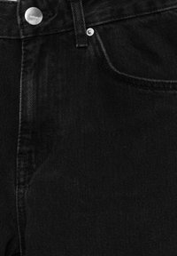 Gros plan sur un jean en denim noir montrant la poche avant, la passants de ceinture, la braguette zippée et un bouton métallique avec le nom de la marque gravé.