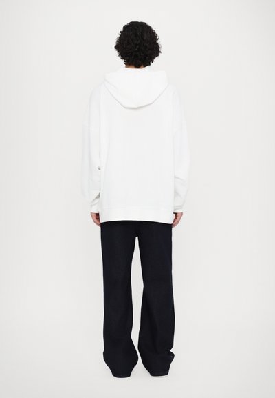 Λευκή κουκούλα με φαρδιά ώμους, συνδυασμένη με σκούρο, φαρδύ παντελόνι. Λείος υφασμάτινος τύπος; χαλαρή εφαρμογή. Απλός, μινιμαλιστικός σχεδιασμός.