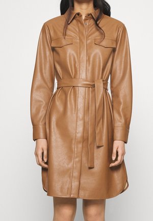 Femme portant une robe-chemise en similicuir beige ceinturée, à manches longues et poches à rabat sur la poitrine, debout devant un fond uni.