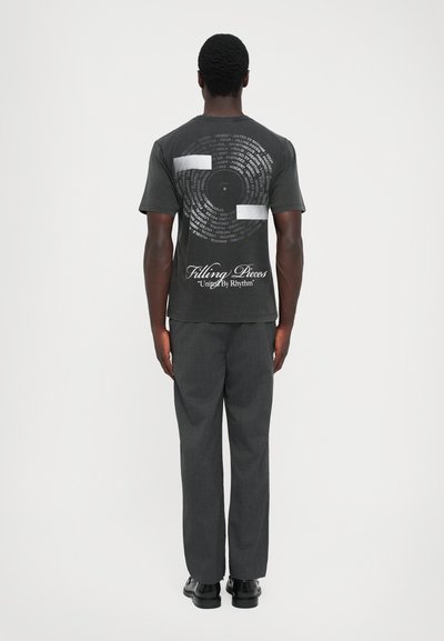 Bărbat în picioare, văzut din spate, purtând un tricou grafic negru cu text circular și sloganul "Filling Pieces", pantaloni gri închis și pantofi negri.