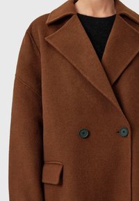 Cappotto doppio petto in misto lana marrone con colletto a rever, grandi bottoni neri e tasca laterale. Texture morbida e silhouette strutturata.
