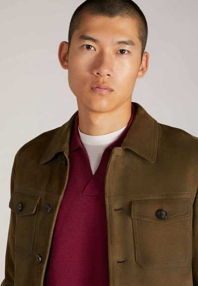 Giacca in suede marrone con tasche sul petto con bottone, abbinata a un maglione a V di colore rosso scuro. Il modello ha i capelli corti e un'espressione neutra.