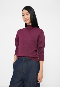 Suéter de gola alta na cor burdigala, feito de um material suave, com mangas longas, um design justo e punhos canelados, combinado com calças azul-escuro.