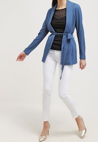 Blauwe, lichte wikkeljas met een strikceintuur, gedragen over een zwarte, gestructureerde top. Gepaard met witte slim-fit broek en metalen hakken.