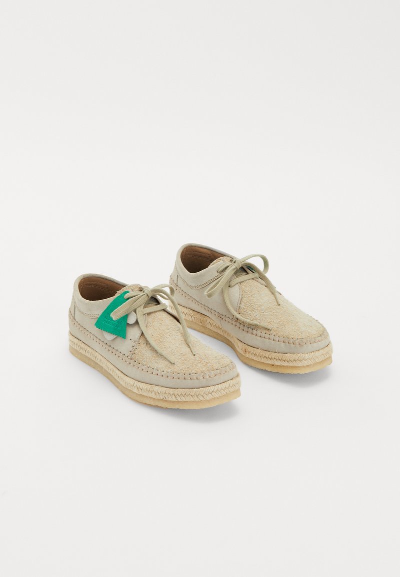 Beige schoenen met een gestructureerde bovenkant, voorzien van een groene tag, platte veters en een geweven touwzool. Ronde neus en stikdetails.