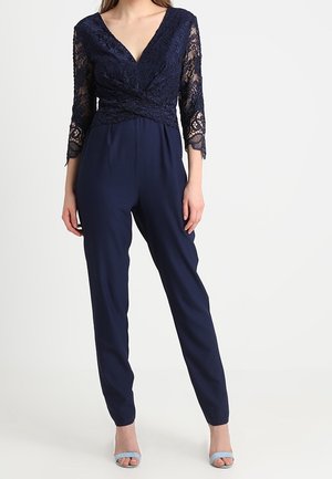 Vrouw draagt een marineblauwe jumpsuit met kanten driekwartmouwen en een V-hals, gecombineerd met lichtblauwe open-toe hakken.