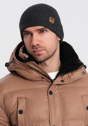 Bruine pufferjack met zwarte accenten en drukknoopsluitingen, gecombineerd met een donkergrijze geribde beanie met een beige logo-patch.