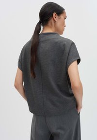 Vrouw met een lange donkere paardenstaart, gekleed in een losse donkergrijze top met korte mouwen en bijpassende grijze broek, staande met haar handen in haar zakken.