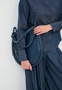 Donkerblauwe leren handtas met een gebogen vorm en knoopdetail op de handgreep. Gladde textuur met gouden hardware accenten.