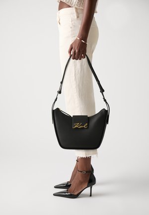 Black leather handbag with a curved design, gold logo detail, and a zip closure. Worn on arm, paired with cream-colored pants and black heels.