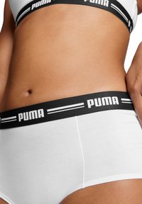Braguita blanca de algodón con una cintura elástica negra que presenta el logo de Puma y dos rayas blancas. Textura suave y diseño simple.