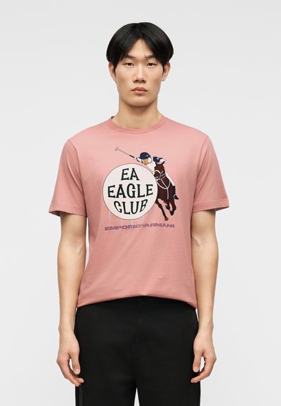 Νέος άνδρας που φοράει ένα ροζ T-shirt με γραφικό παίκτη πόλο και το κείμενο "EA EAGLE CLUB Emporio Armani", συνδυασμένο με μαύρο παντελόνι.