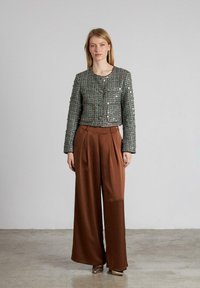 Grüne strukturierte Jacke mit silbernen Knöpfen und Taschen, kombiniert mit hoch taillierten braunen weiten Hosen, vor einem neutralen Hintergrund.