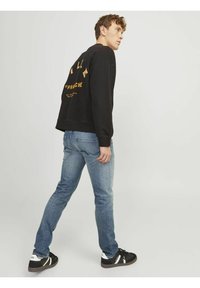 Sweatshirt noir avec des graphismes jaunes au dos, associé à un jean en denim bleu clair et des baskets noires avec des accents blancs. Look décontracté.