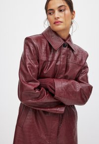 Cappotto in ecopelle bordeaux con un motivo a pelle di coccodrillo texturizzato, caratterizzato da un ampio colletto e bottoni grandi, progettato con maniche oversize.