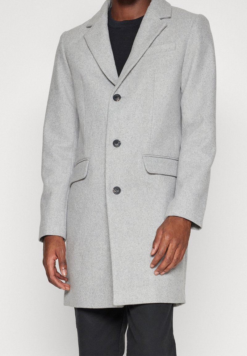 Homme portant un manteau en laine gris clair boutonné par-dessus un pull noir avec un pantalon noir, debout devant un fond uni.