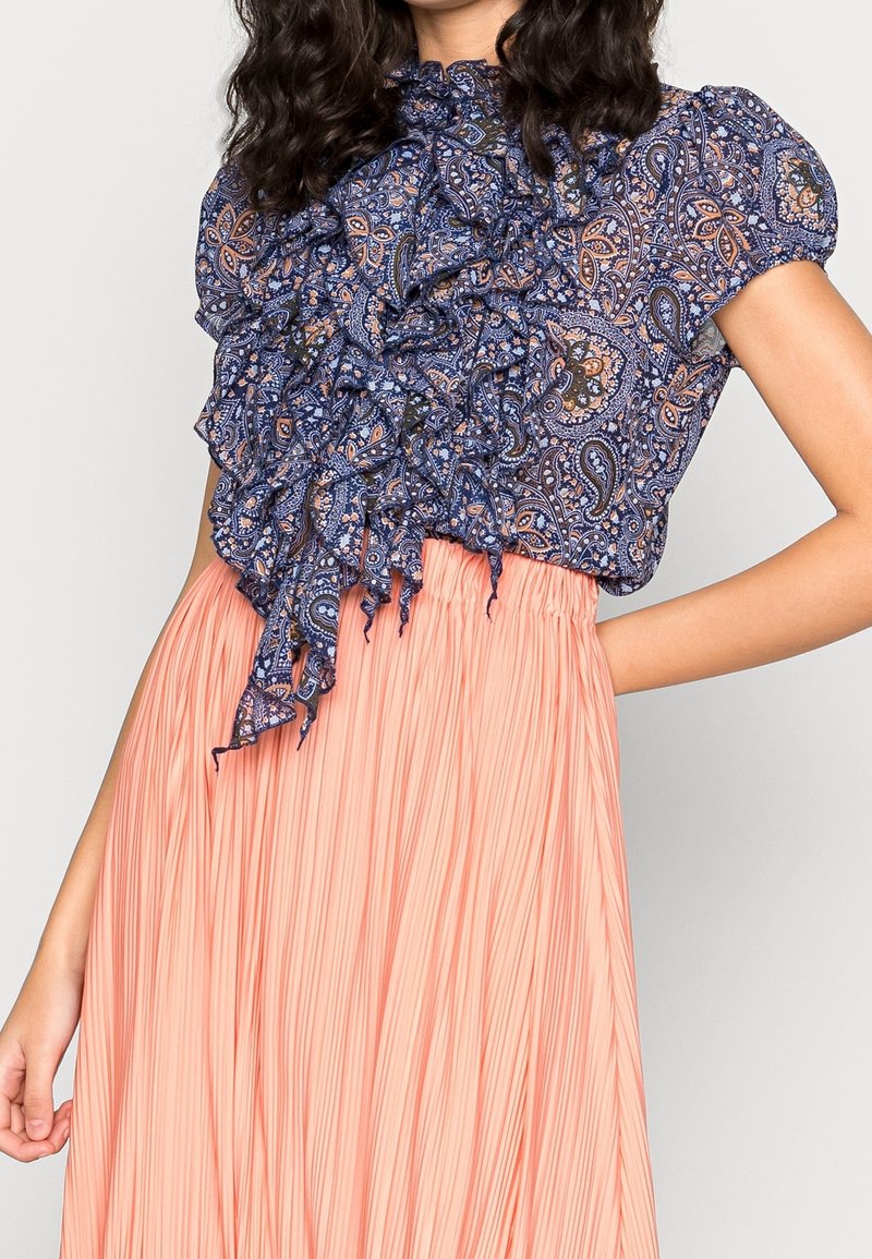 Blusa blu con motivo paisley, con scollatura arricciata e maniche a sbuffo corte; abbinata a una gonna plissettata color pesco. Tessuti morbidi e fluido.