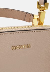 Lys beige lærveske med gullglidelås og metallbeslag. "Coccinelle" er preget i gull på utsiden for merkeidentifikasjon.
