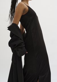 Robe noire sans manches avec une texture lisse et élégante et une fente sur le côté. Le modèle tient un manteau noir ample assorti avec des poignets à boutons.