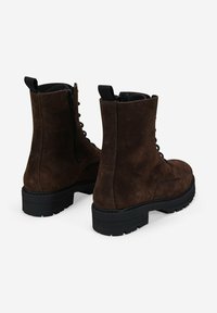 Botas de ante marrón oscuro con cordones, suelas de goma gruesa negra y cremalleras laterales, mostradas desde atrás y con una vista lateral ligeramente angulada sobre un fondo blanco.