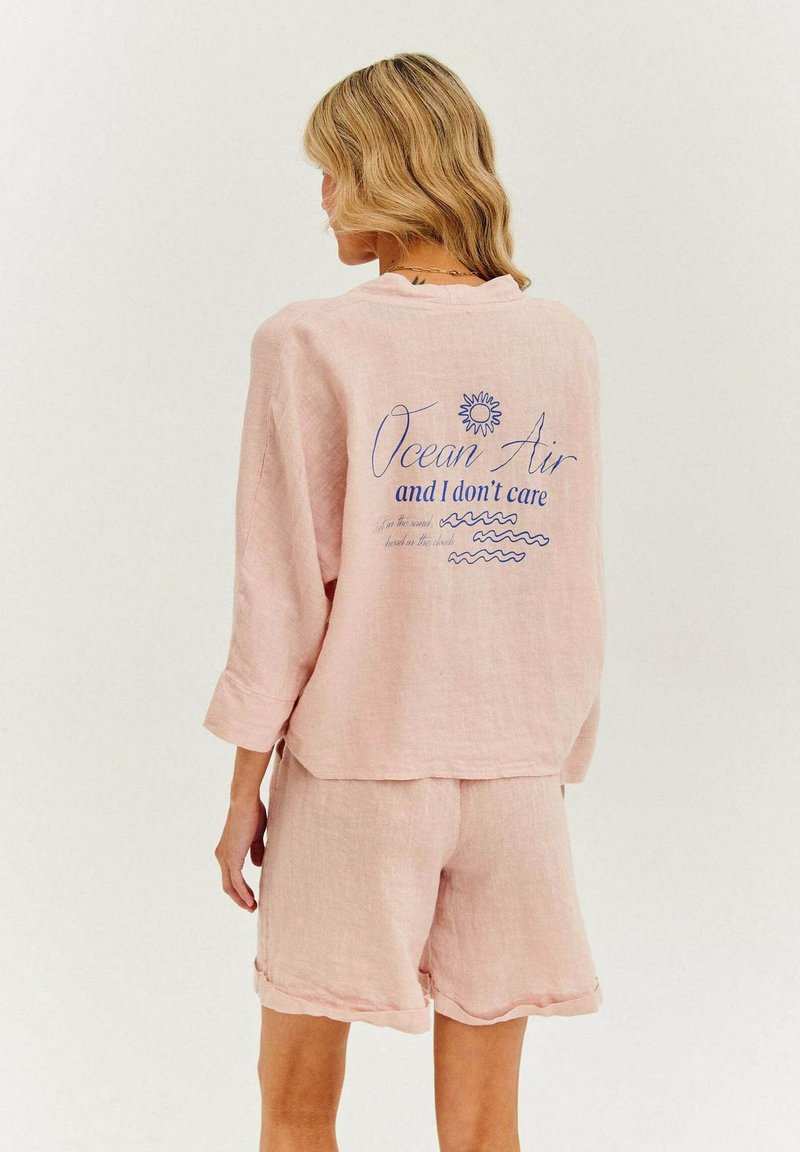 Svetloružový ľanový top s dlhými rukávmi, so modrým textom a grafikou slnka. Skombinovaný s rovnakými ružovými šortkami s vykrojeným lemom.