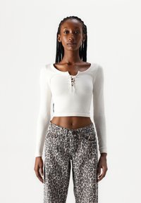 Fehér, hosszú ujjú, bordás crop top, amelynek mély kivágása és fűzős részlete van, magasderekú, leopárdmintás farmernadrággal párosítva.