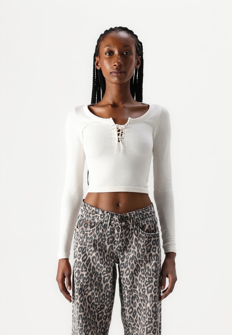 Fehér, hosszú ujjú, bordás crop top, amelynek mély kivágása és fűzős részlete van, magasderekú, leopárdmintás farmernadrággal párosítva.