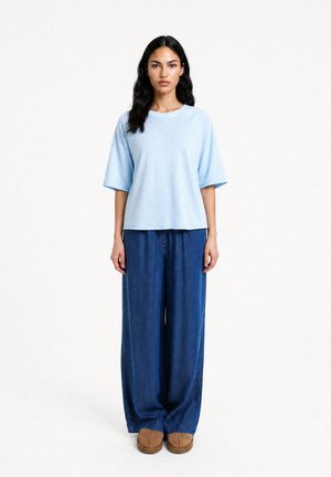 Frau mit langen dunklen Haaren, die ein hellblaues übergroßes T-Shirt, weite blaue Jeans und braune Schuhe vor einem einfarbigen weißen Hintergrund trägt.