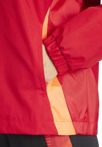 Veste coupe-vent rouge avec des poignets élastiques, dotée d'une poche latérale doublée de tissu orange, texture lisse et matériau léger.